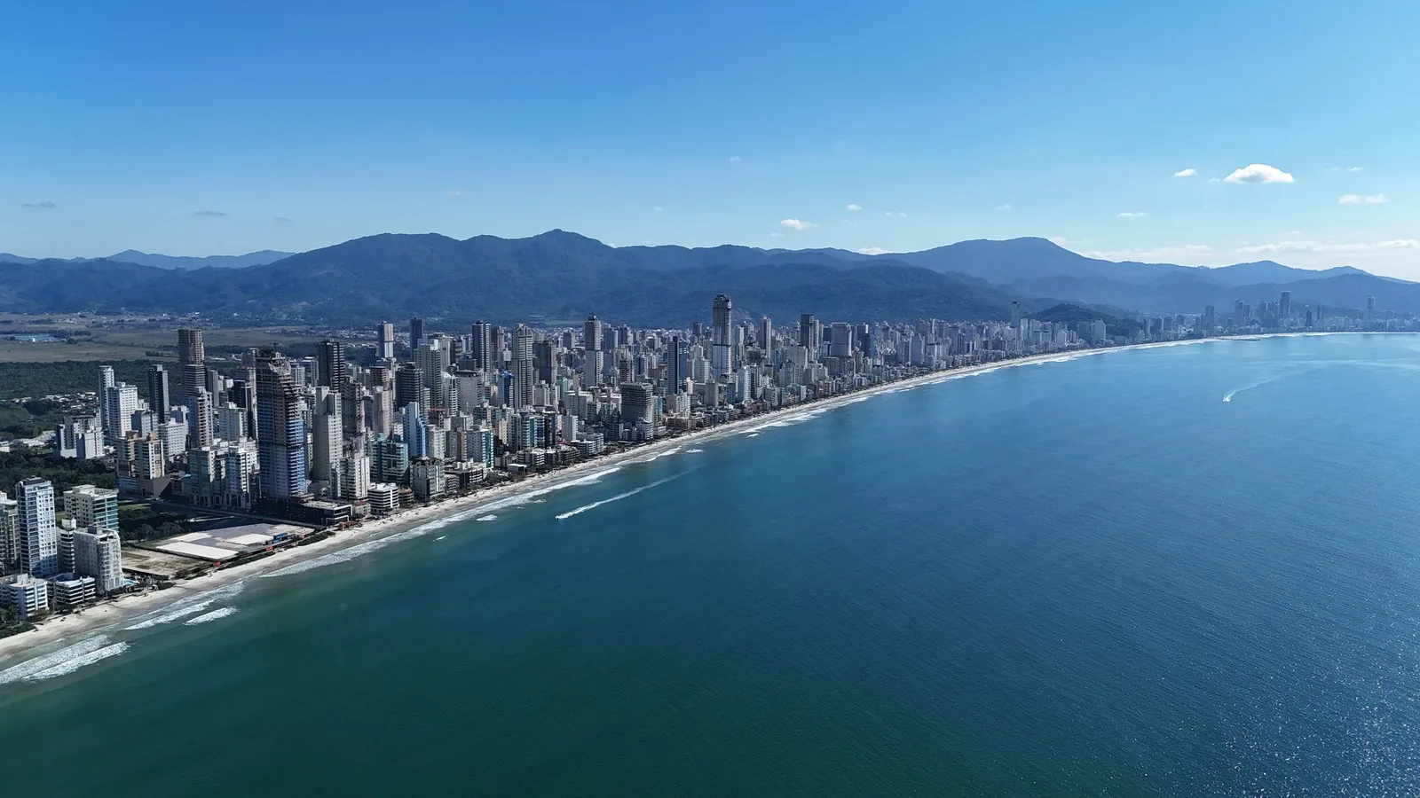 Panorama aéreo da Meia Praia, em Itapema, com uma linha contínua de prédios altos ao longo da praia, mar azul e serra verde ao fundo.