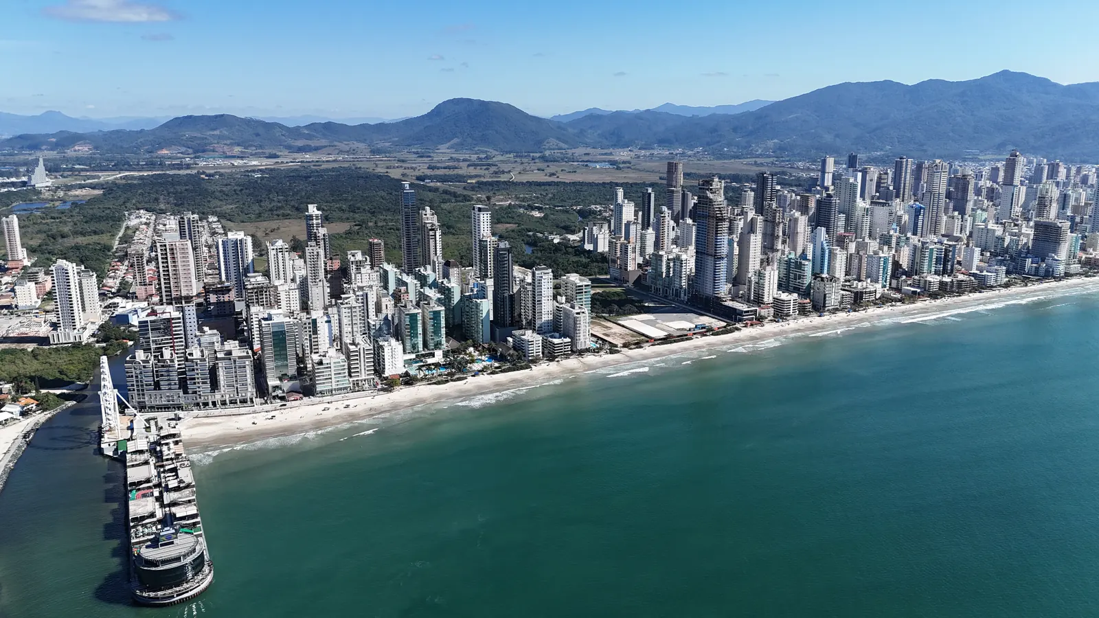 Vista aérea da Meia Praia, em Itapema, com prédios altos de frente para o mar, a faixa de areia, o píer no canto e montanhas ao fundo.